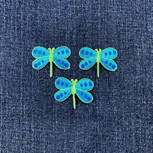 May include: Three embroidered dragonfly patches. Each dragonfly has blue wings with darker blue dot patterns and a green body. The patches are on a blue denim background.