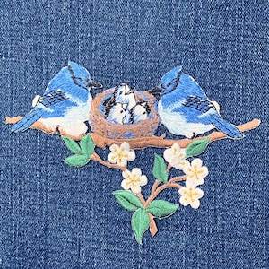 May include: Embroidered patch featuring two blue birds perched on a branch with a nest containing baby birds. The patch includes white flowers and green leaves, all against a blue denim background.
