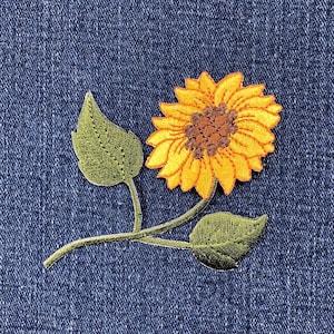 May include: Embroidered sunflower patch with yellow petals and a brown center, accompanied by green leaves and stems. The patch is set against a blue denim background, showcasing the vibrant colors and detailed stitching.