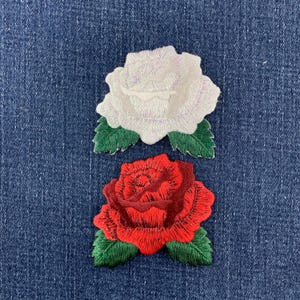 May include: Two embroidered rose patches featuring detailed stitching. The top patch is a white rose with a subtle iridescent sheen, and the bottom patch is a vibrant red rose. Both flowers are accented with dark green leaves at the base.