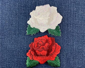 May include: Two embroidered rose patches featuring detailed stitching. The top patch is a white rose with a subtle iridescent sheen, and the bottom patch is a vibrant red rose. Both flowers are accented with dark green leaves at the base.