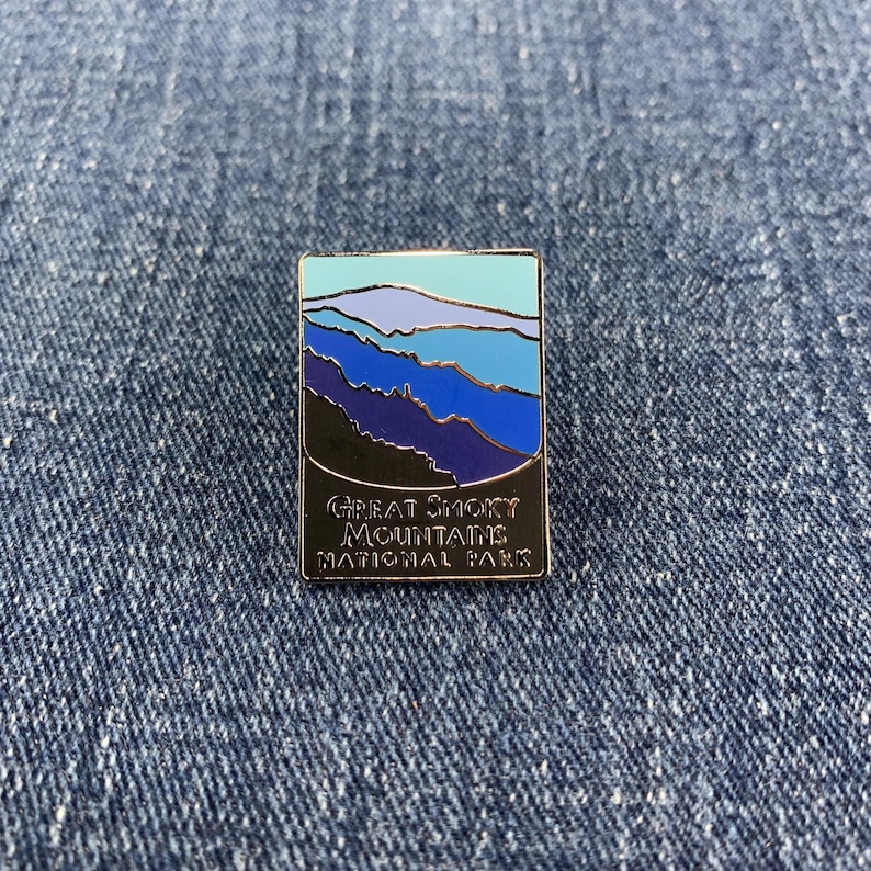 May include: Rectangular enamel pin featuring a landscape design of the Great Smoky Mountains National Park. The pin has a black border and the text "Great Smoky Mountains National Park". The landscape design includes shades of blue, purple, and teal.