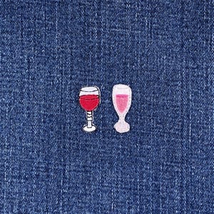 May include: Two embroidered wine glass patches on a blue denim background. One glass is filled with red wine, the other with pink wine. Both patches have white outlines and are detailed with delicate stitching.