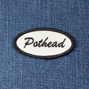 May include: White oval patch with black stitching and the word "Pothead" written in a black cursive font.
