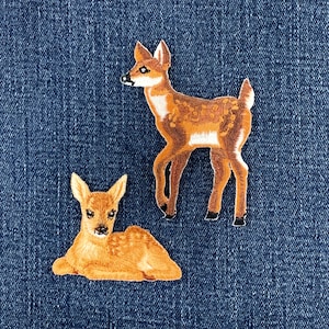 May include: Two embroidered fabric patches featuring deer. One patch depicts a standing fawn with brown fur, white spots, and a white underbelly. The second patch shows a fawn lying down with similar brown fur and white spots on its back.