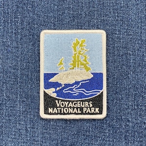 May include: A blue and white embroidered patch with a green tree and the text "Voyageurs National Park".