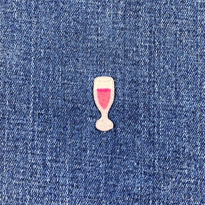 Könnte beinhalten: Ein kleiner, gestickter Aufnäher in Form eines Sektglases. Das Glas ist weiß mit einer rosa Flüssigkeit im Inneren. Der Aufnäher befindet sich auf einem blauen Jeanshintergrund.