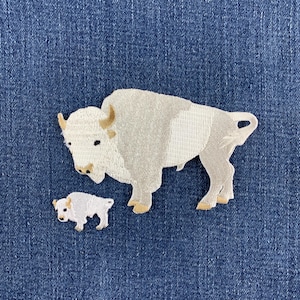 May include: An embroidered patch featuring a large white bison and a smaller white bison calf. The bison are depicted in shades of white and light gray with tan accents on the horns and hooves, set against a textured dark blue denim fabric background.