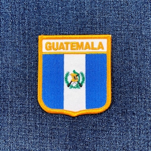 May include: Embroidered patch featuring the Guatemalan flag. The patch has a shield shape with a blue and white striped background, a central emblem, and the word "GUATEMALA" in gold lettering. The patch is outlined in gold and set against a blue denim background.