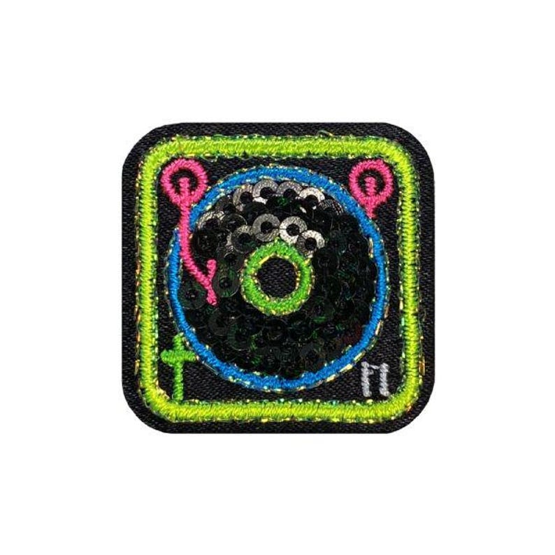 May include: A square, embroidered patch featuring a stylized record player design. The record is black with silver sequins, surrounded by a blue ring. The patch has a neon green border with pink and yellow accents.