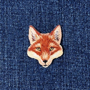 May include: Embroidered patch featuring a fox head with orange and cream-colored fur and yellow eyes. The patch is set against a blue denim background, showcasing the detailed stitching and design of the animal.