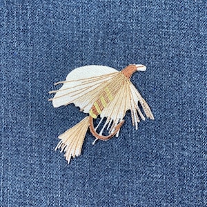 May include: A brown and white embroidered fly fishing lure patch. The lure is on a blue denim background.