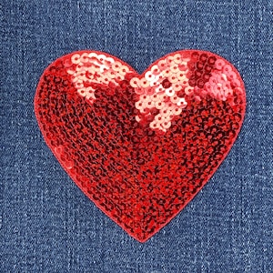 May include: A close-up of a red sequined heart-shaped patch on a blue denim background. The heart is filled with small, shiny red sequins. The denim has a textured appearance.