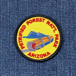 May include: Embroidered patch featuring a yellow circle with the text "PETRIFIED FOREST NATL PARK ARIZONA" in red. The patch has a black border and depicts a stylized illustration of a log and a blue structure.