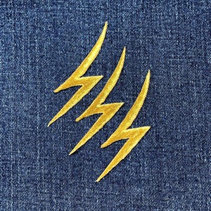 May include: Close-up of a blue denim fabric with three embroidered gold lightning bolt patches. The bolts are arranged diagonally across the frame, adding a dynamic visual element to the design.