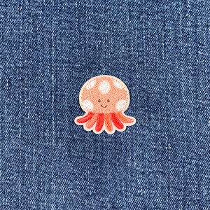 May include: A small, embroidered patch featuring a cartoon jellyfish design. The jellyfish has a peach-colored head with white polka dots and a smiling face. Its tentacles are red-orange. The patch is likely for clothing or accessories.