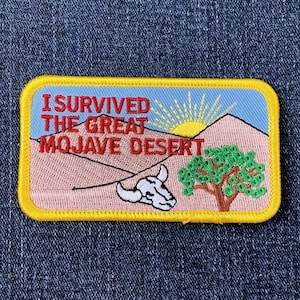 Op de afbeelding: Geborduurde patch met de tekst "I Survived The Great Mojave Desert". De patch toont een woestijnlandschap met een zon, bergen, een boom en een koeienschedel. De patch heeft een gele rand.