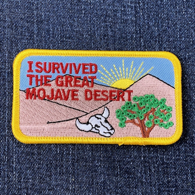Op de afbeelding: Geborduurde patch met de tekst "I Survived The Great Mojave Desert". De patch toont een woestijnlandschap met een zon, bergen, een boom en een koeienschedel. De patch heeft een gele rand.