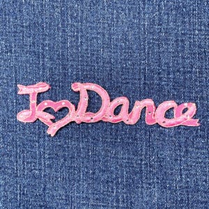 Peut inclure: Un écusson brodé rose avec les mots "I ♡ Dance" dans une police cursive. Le symbole du cœur remplace le mot "love". L'écusson est orné de petits strass scintillants et est posé sur un fond en denim bleu.