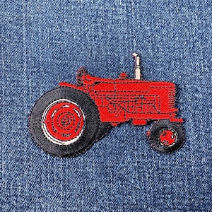 May include: A red tractor patch with black tires and a white accent on a blue denim background. The tractor has a detailed embroidered design, showcasing its wheels, body, and exhaust pipe. This patch is suitable for clothing or accessories.