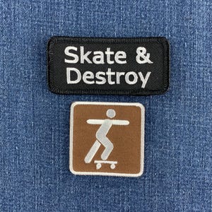 May include: A rectangular black embroidered patch with white text reading Skate & Destroy and a square brown embroidered patch featuring a white graphic of a person on a skateboard, both displayed on a blue denim fabric background.