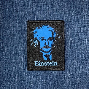 May include: A black rectangular patch with a blue image of Albert Einstein. The image is a stylized portrait of Einstein's face and hair, with the name 'Einstein' below. The patch is likely fabric and is set against a blue denim background.