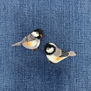 May include: Two embroidered bird patches featuring black heads, white cheeks, grey wings with black detailing, and pale yellow underbellies. The birds are depicted in profile, showcasing detailed thread work on a textured blue denim fabric background.