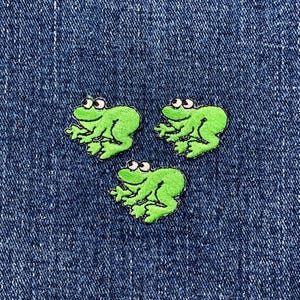 May include: Three embroidered green frog patches with white eyes are arranged on a blue denim background. The frog patches are a bright, cheerful green color and have a cartoon-like design.
