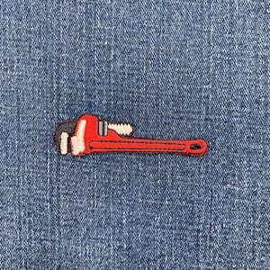May include: A red and black embroidered patch in the shape of a pipe wrench. The patch is on a blue denim background. The wrench has a black handle and a red body.