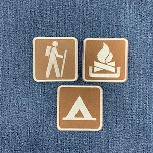 May include: Three square embroidered patches with rounded corners, featuring a tan background and white stitching. The designs depict a person hiking with a backpack, a campfire with logs, and a triangular tent, all outlined in white thread.