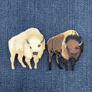May include: Two embroidered patches featuring bison figures. One patch depicts a cream-colored bison with light brown accents, while the other patch shows a brown bison with a dark brown head, mane, and legs, both rendered in detailed thread work.