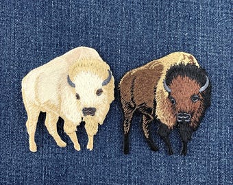 May include: Two embroidered patches featuring bison figures. One patch depicts a cream-colored bison with light brown accents, while the other patch shows a brown bison with a dark brown head, mane, and legs, both rendered in detailed thread work.