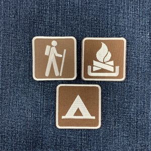 May include: Three square embroidered patches with rounded corners, featuring a brown background and white stitched icons. The icons depict a person hiking with a backpack, a campfire with logs, and a triangular tent, all with white borders.