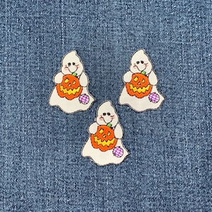 Könnte beinhalten: Drei weiße, geisterförmige Stoff-Patches mit gestickten Details. Jeder Geist hält einen kleinen orangefarbenen Kürbis mit einem Jack-o'-Lantern-Gesicht. Die Geister haben lila und weiße Tupfen-Akzente.
