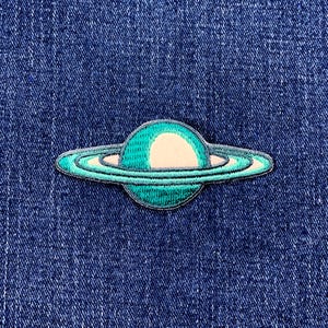 May include: An embroidered patch featuring a teal planet with a cream-colored center and a ring system. The patch is stitched with teal and white thread, and is set against a blue denim background.