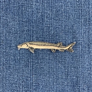 Könnte beinhalten: Ein gestickter Aufnäher in Form eines Störs. Der Fisch ist in Beige-, Hellbraun- und Dunkelbrauntönen gehalten und zeigt einen langen, schlanken Körper, eine spitze Schnauze, sichtbare Flossen und eine gegabelte Schwanzflosse mit Details.