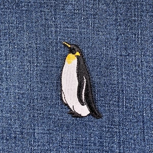 May include: Embroidered penguin patch in black, white, and yellow. The penguin is depicted standing on a blue denim background. This patch is likely designed for application on clothing or accessories.