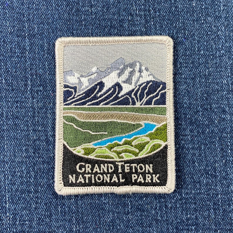 May include: A white embroidered patch with a blue river winding through a green valley. The mountains in the background are white with a gray sky. The text on the patch reads 'Grand Teton National Park'.