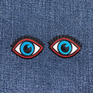 May include: Two embroidered eye patches. Each patch features a blue iris, black pupil, white sclera, and a red outline. The patches are bordered by black, bead-like embellishments. The patches are set against a blue denim background.
