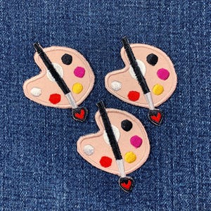 May include: Three embroidered artist palette patches. Each patch features a light pink palette with colorful paint splotches in red, pink, yellow, black, and white. A black paintbrush with a red heart-shaped tip is included.