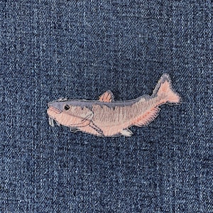 May include: Embroidered patch featuring a fish design in shades of pink, tan, and gray, with a white whisker detail. The patch is set against a textured, dark blue denim background, creating a contrast in color and texture.