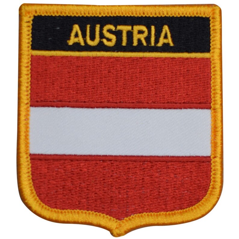 May include: Embroidered patch featuring the Austrian flag design. The patch has a shield shape with a gold border. The flag design includes red and white horizontal stripes, with the word "AUSTRIA" in gold letters on a black background.