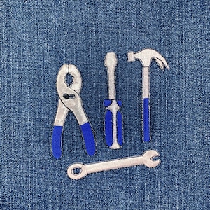 May include: Embroidered tools including pliers, a screwdriver, a hammer, and a wrench, with blue and silver accents, are arranged on a blue denim background. The tools are depicted in a simple, illustrative style, suitable for a craft or DIY theme.