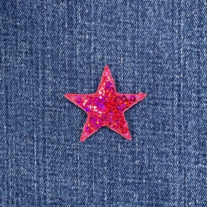 May include: A close-up of a red sequined star on a blue denim background. The star is a vibrant shade of red with a glittery texture, and it is set against the textured denim fabric.