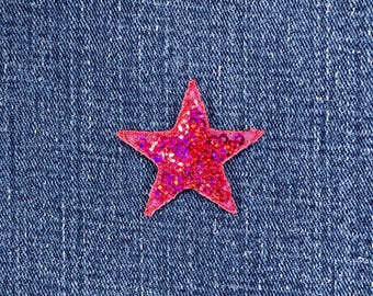May include: A close-up of a red sequined star on a blue denim background. The star is a vibrant shade of red with a glittery texture, and it is set against the textured denim fabric.