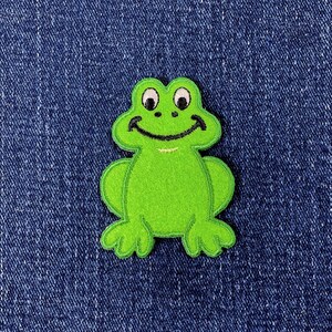 May include: A bright lime green embroidered patch in the shape of a cartoon frog. The frog features two white eyes with black pupils, two small black nostrils, and a wide black stitched smile. The edges are finished with a matching green border.