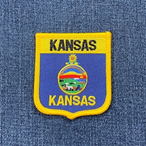 May include: A blue and yellow embroidered patch featuring the state seal of Kansas. The seal depicts a sunflower, a river, and a field of wheat. The words "Kansas" are printed in black at the top and bottom of the patch.