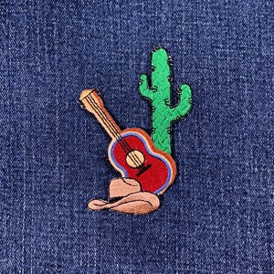 May include: Embroidered patch featuring a red guitar with a blue outline, a green cactus, and a brown cowboy hat. The patch is set against a blue denim background, evoking a Western or country music theme.