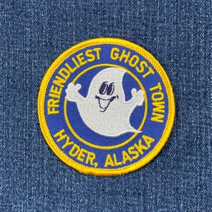 Könnte beinhalten: Ein gestickter Aufnäher in Gelb und Blau mit dem Text "Friendliest Ghost Town Hyder, Alaska". In der Mitte des Aufnähers befindet sich ein weißer Geist mit einem lächelnden Gesicht.