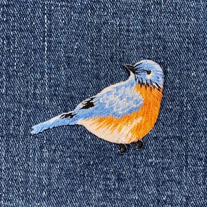 Pu&ograve; includere: Toppa ricamata di un uccello azzurro con dettagli blu, arancioni e neri. L'uccello &egrave; cucito su uno sfondo di denim. La toppa &egrave; un oggetto decorativo, adatto per abbigliamento o accessori.